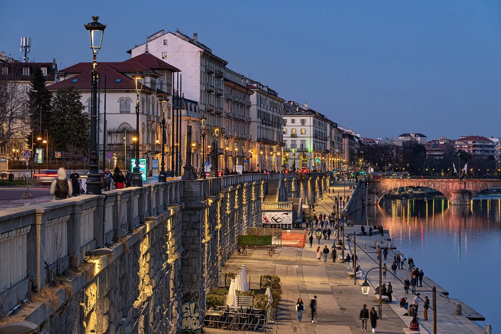 I Murazzi del Po a Torino all'imbrunire, con la banchina illuminata lungo il fiume, i palazzi sopra gli argini e un ponte in pietra sullo sfondo