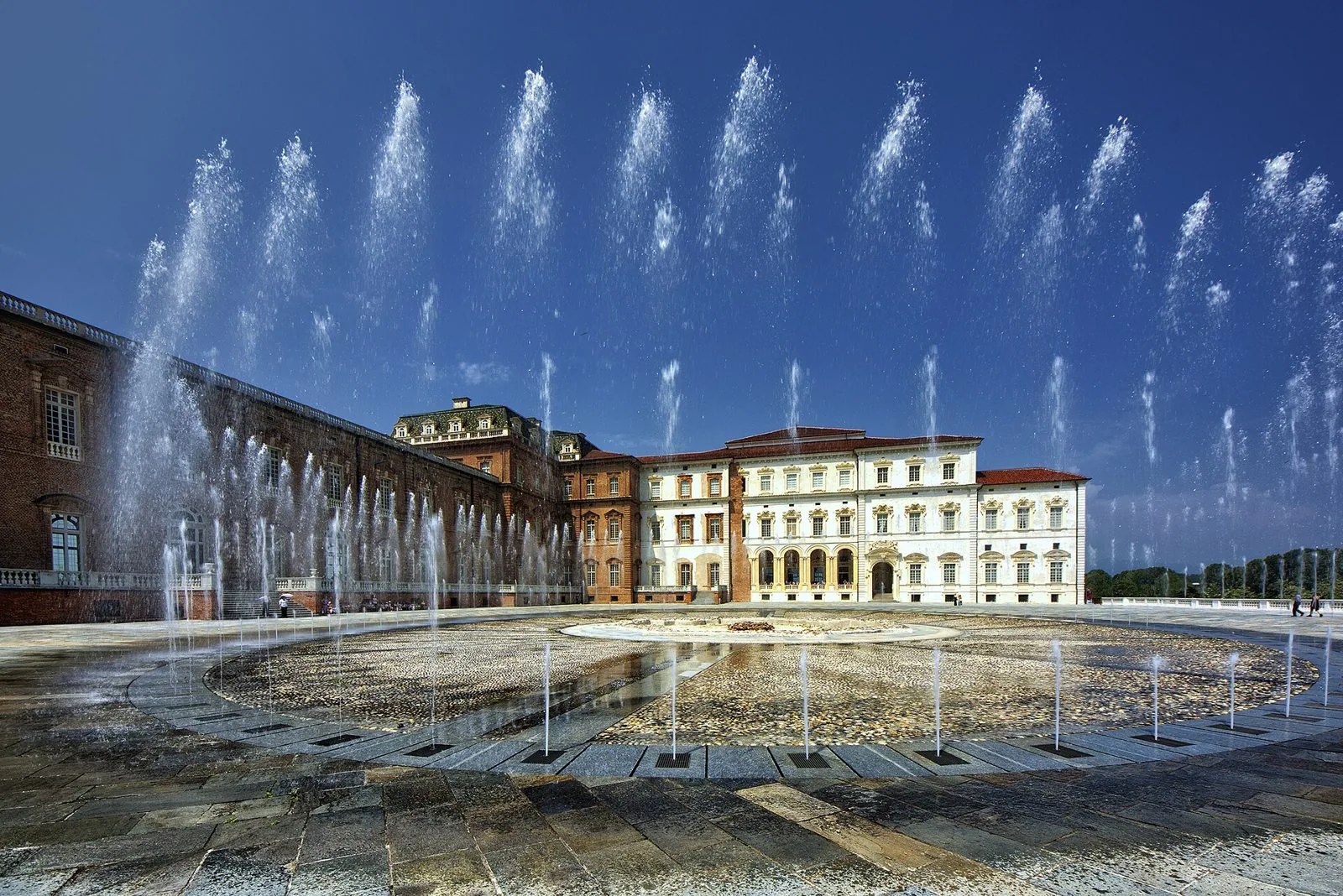 La corte d'onore della Reggia di Venaria Reale con i getti d'acqua della fontana in primo piano e le facciate barocche sotto un cielo blu