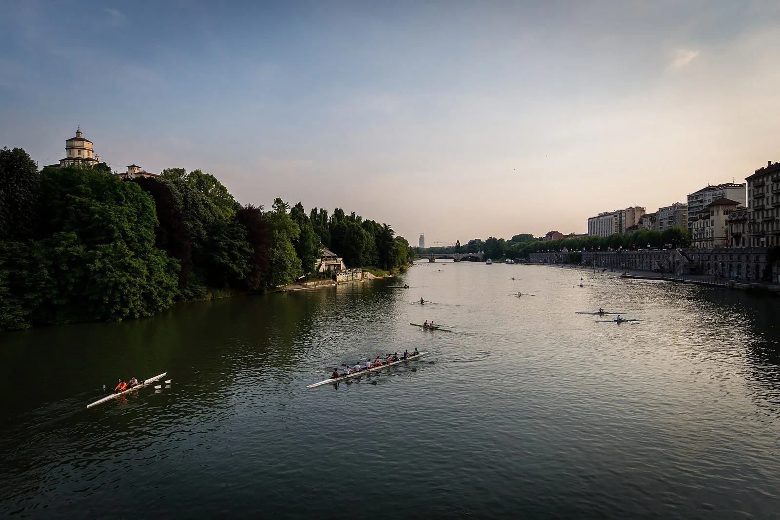 Il Po a Torino al tramonto con barche di canottieri, la chiesa del Monte dei Cappuccini sulla collina a sinistra, un ponte sullo sfondo e i Murazzi a destra