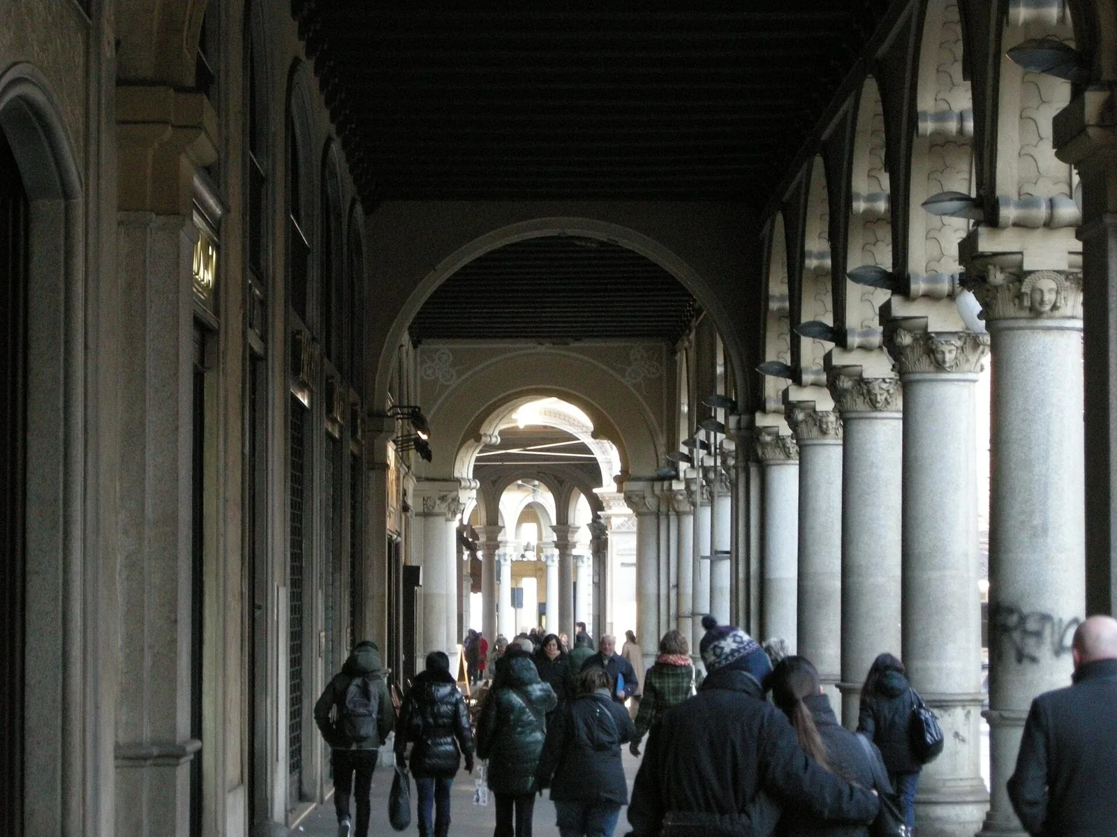 Passanti in abiti invernali camminano sotto i portici di via Pietro Micca a Torino, tra colonne in pietra e arcate